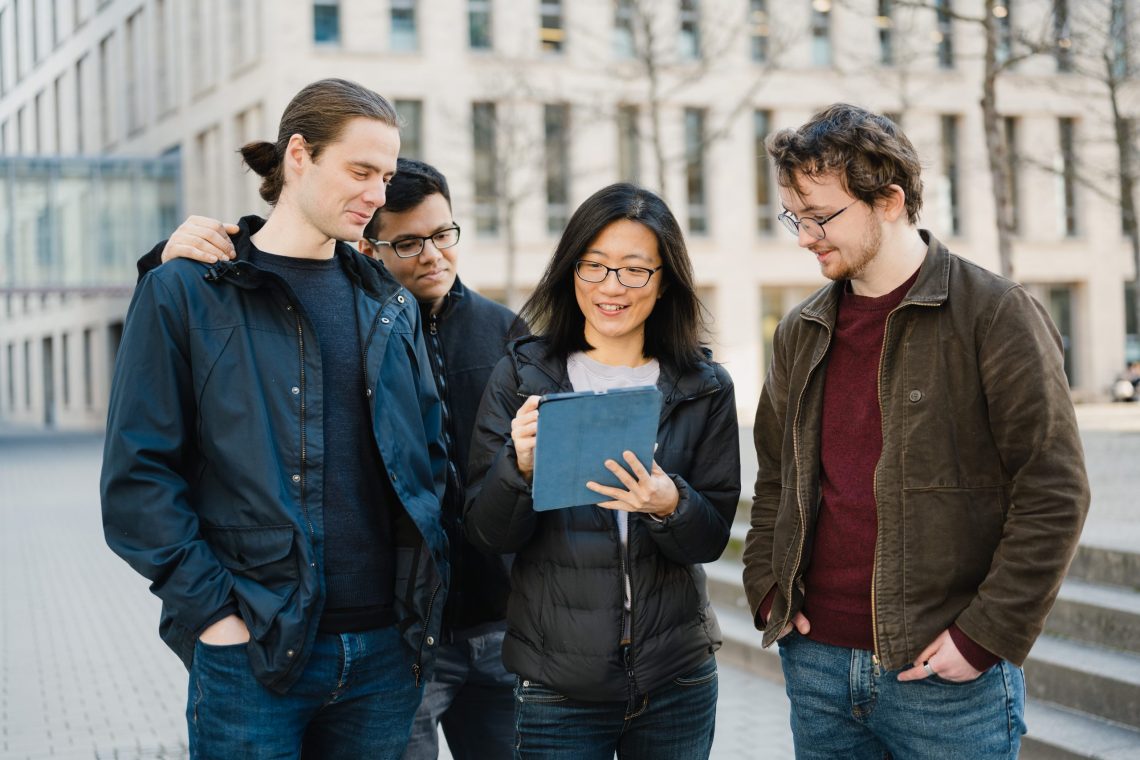 Vier verschiedene Studierende arbeiten gemeinsam an einem Tablet vor einem Universitätsgebäude und demonstrieren Teamarbeit in einer akademischen Umgebung
