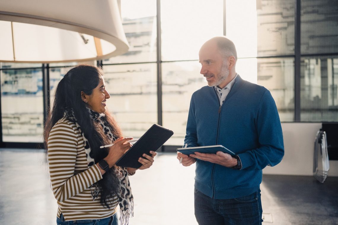 A woman and a man are standing in a modern, sunlit building having a conversation. Both are holding digital tablets and styluses, appearing to discuss something work-related. The woman is smiling and wearing a striped sweater with a scarf, while the man wears a blue zip-up jacket. Large windows in the background allow natural light to fill the space.