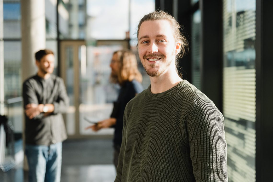 Smiling young man with long hair and beard wearing a green sweater in a bright office space, with colleagues conversing in the background.