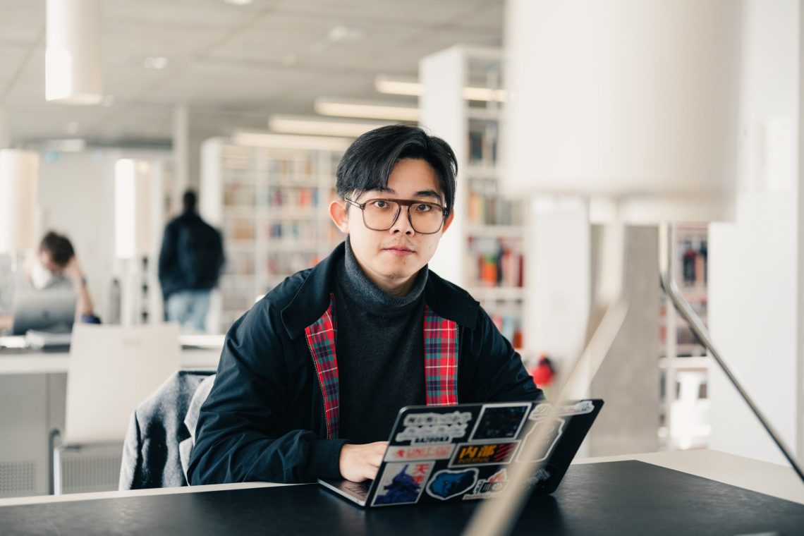 A young man sits at a desk in a bright, modern library, looking directly at the camera. He is using a laptop covered in various stickers and wears glasses, a dark turtleneck, and a jacket with a red tartan lining. In the background, other people are working or walking among shelves filled with books.