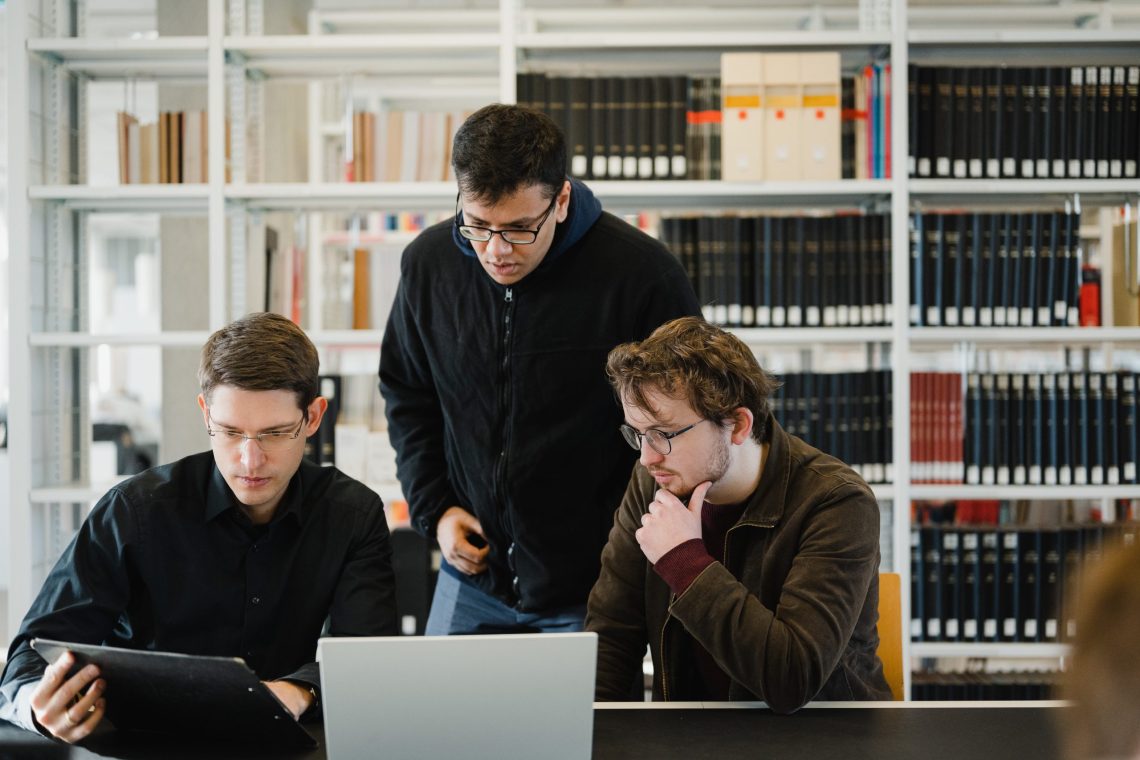 Three young men are working together at a table in a library. One of them is showing something on a tablet, while another is looking at a laptop screen thoughtfully with his hand on his chin. The third man is standing and leaning in to observe. Shelves filled with books and files are visible in the background.