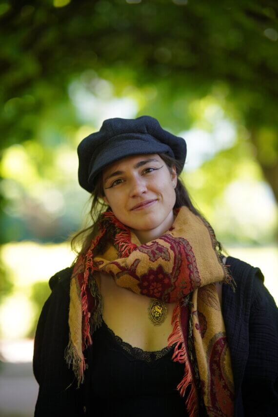 Person wearing a dark outfit, patterned scarf, and beret standing outdoors under leafy green trees