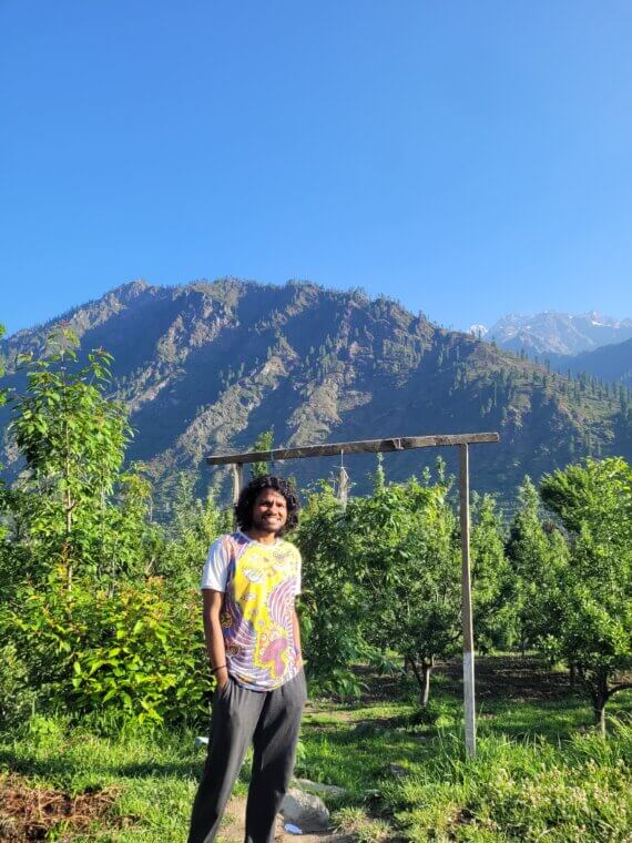 Person standing in a green orchard with tall mountains and a clear blue sky in the background.